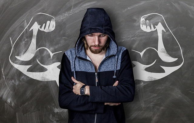 a man wearing a hoodie with arms crossed and staring down, stands in front of a blackboard where a chalk drawing of a mans muscled arms are behind