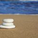 Three white beach pebbles balanced by graduated size on a brown sandy beach with a blue ocean in the background